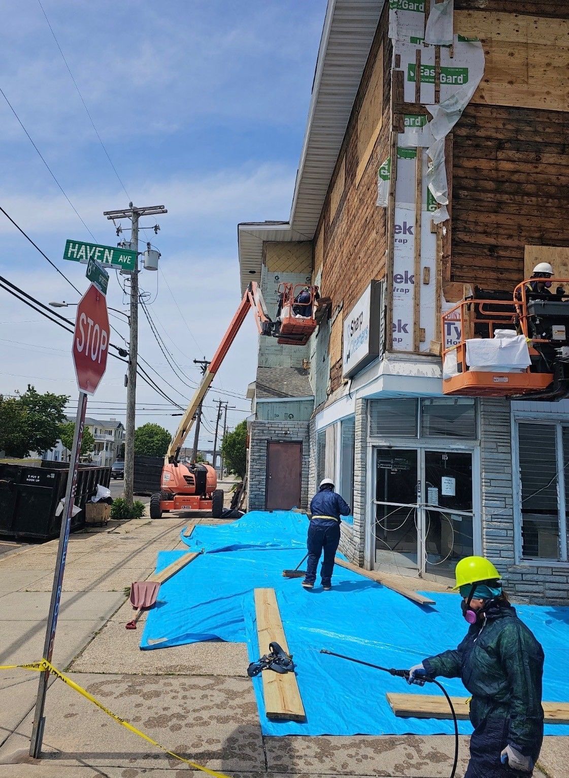 Workers securing a bright blue tarp on a sidewalk beside a building, with a lift and utility poles nearby.