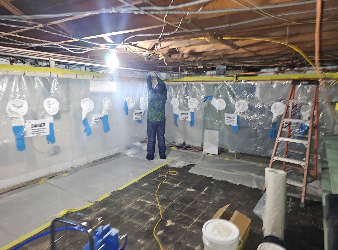 Person spray-painting a basement ceiling with plastic-covered walls and a ladder nearby