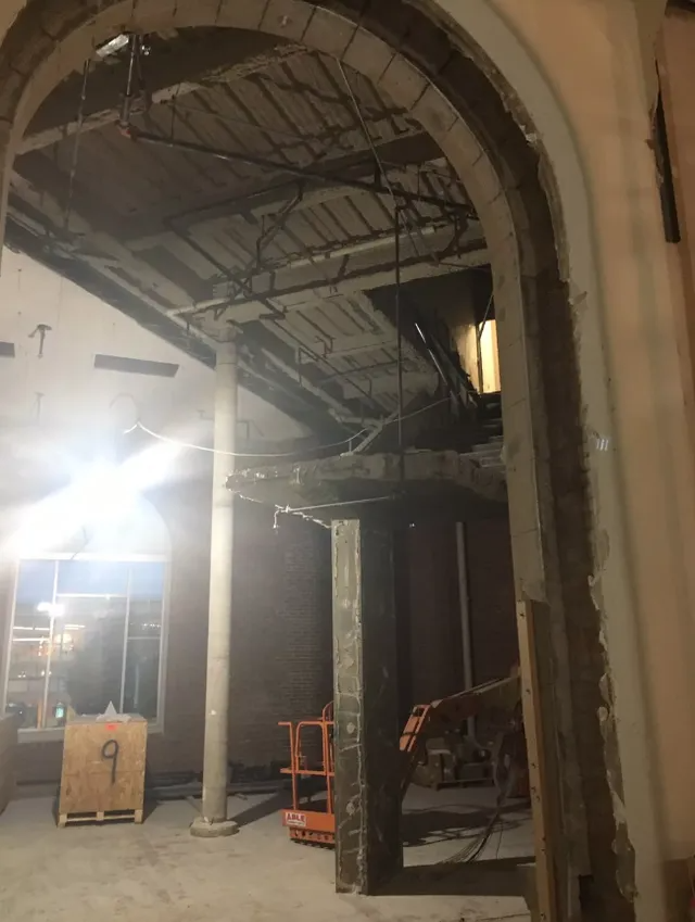 Partially demolished interior with exposed ceiling beams, archway, scaffolding, and construction equipment under bright light