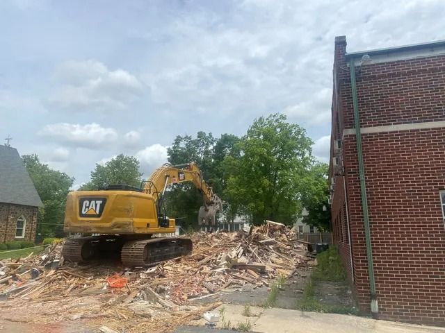 Yellow CAT excavator demolishing a brick building, with rubble piled beside it under a cloudy sky.