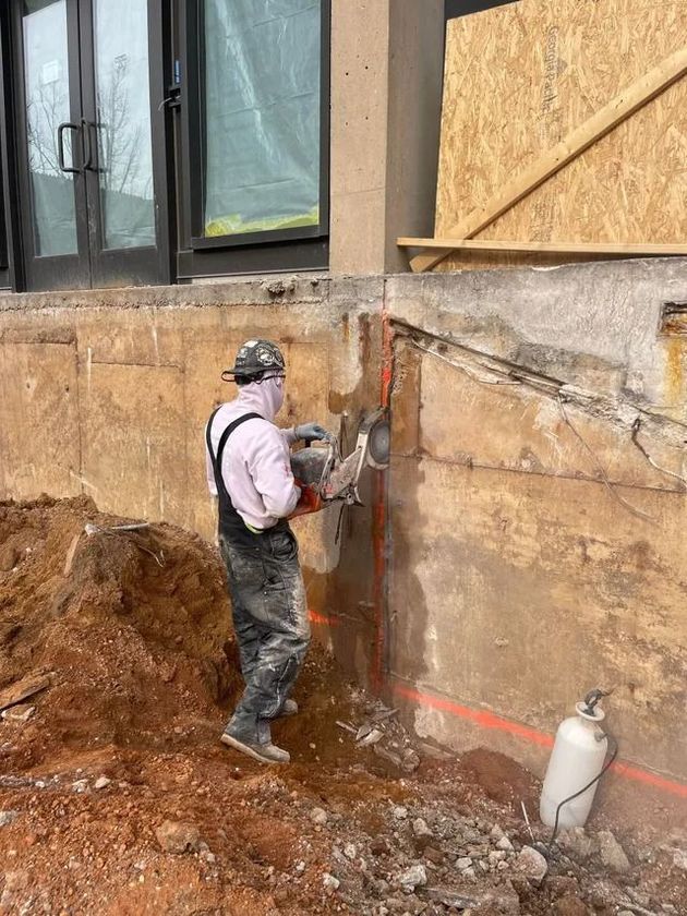 Worker using a jackhammer to break concrete foundation wall beside a house during excavation