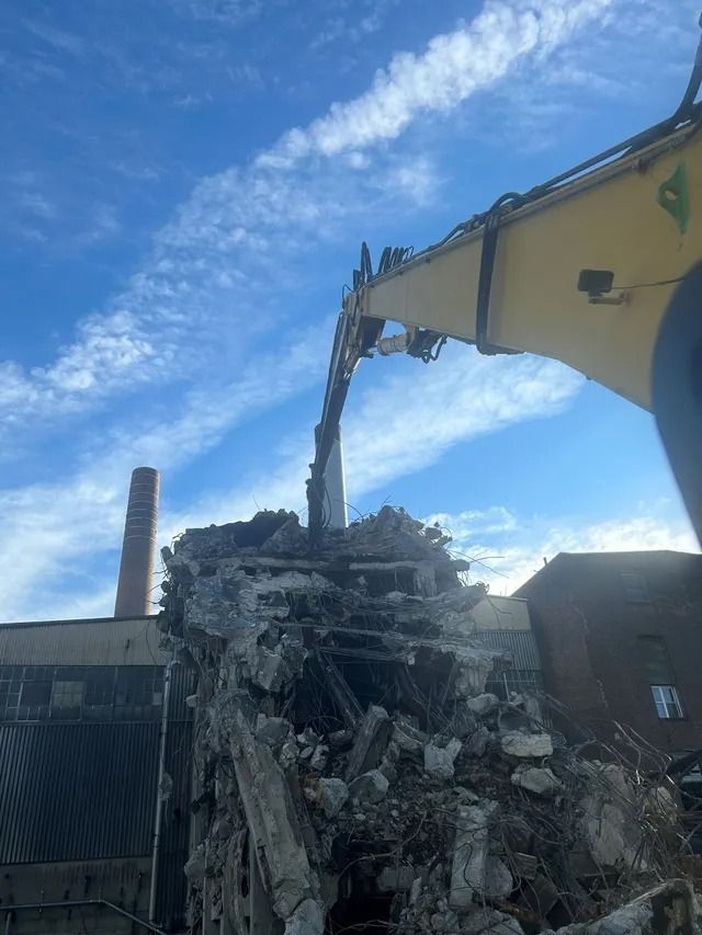 Excavator demolishing a brick wall beside an industrial building under a blue sky
