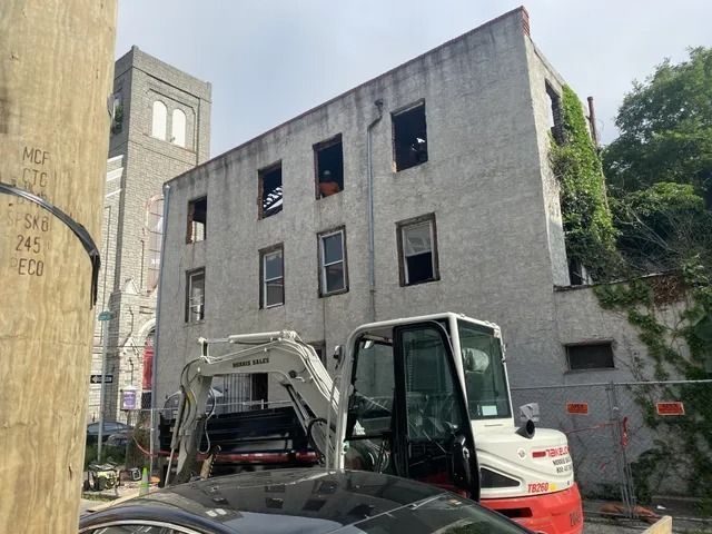 Small excavator beside a gray three-story building with open windows and a red vehicle in front