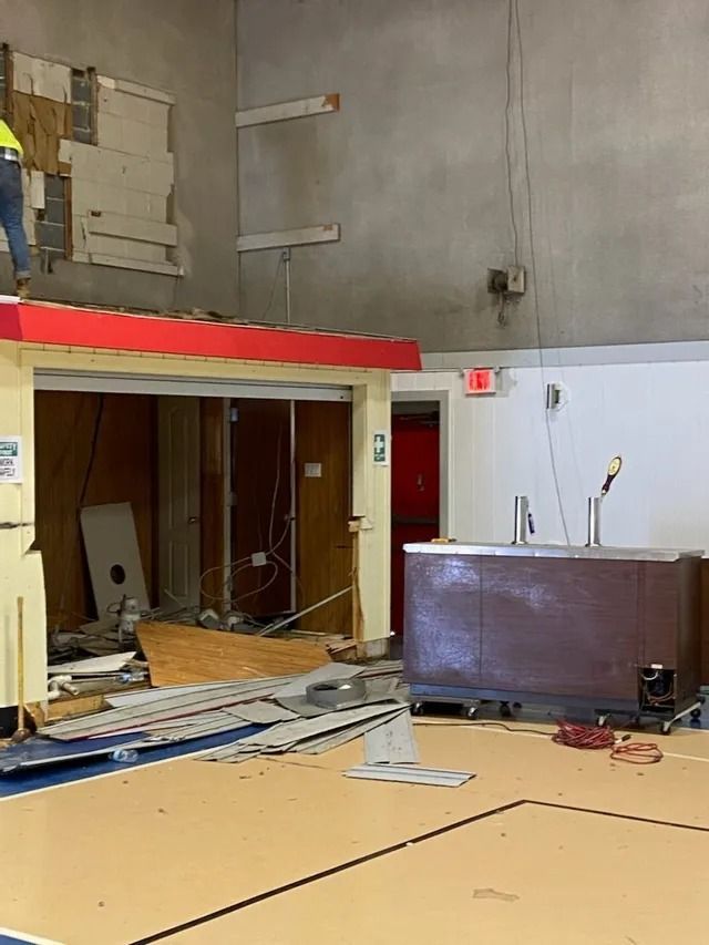 Damaged gym interior with broken wall sections, debris on the floor, and a damaged scoreboard desk