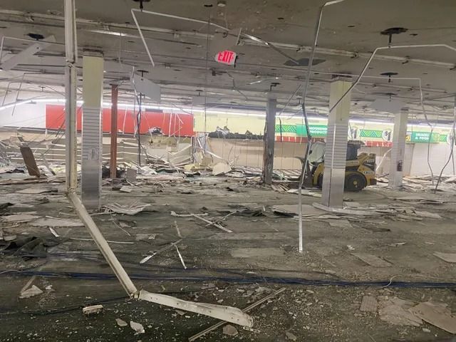 Severely damaged store interior with debris, broken ceiling tiles, and a forklift amid rubble.