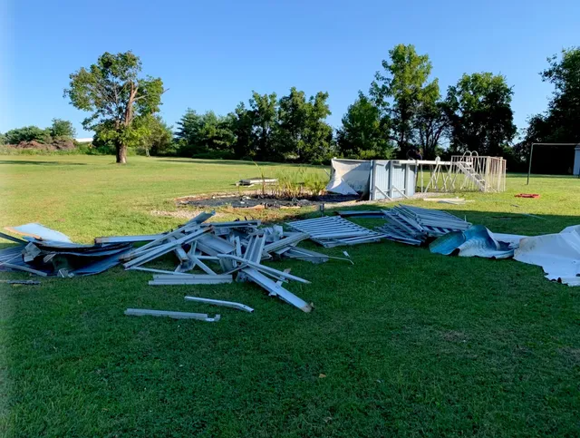Collapsed white metal frame debris on a grassy field with trees and a small structure in the background
