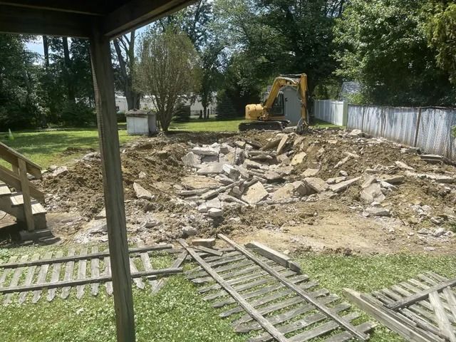 Backyard demolition with an excavator clearing broken concrete and debris near a wooden fence