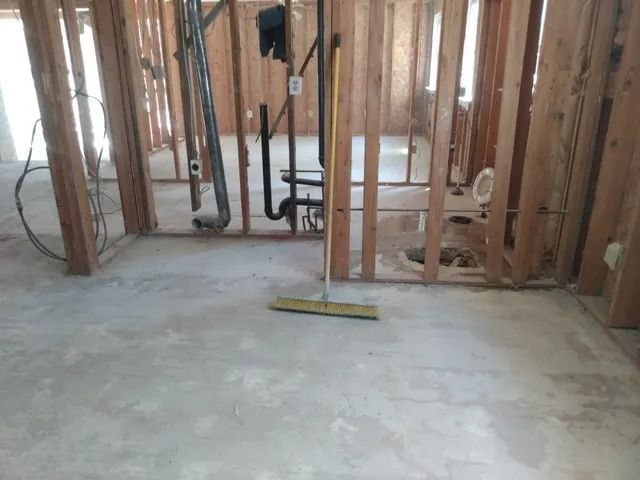 Unfinished room with exposed wall studs, plumbing, and a broom on a concrete floor.