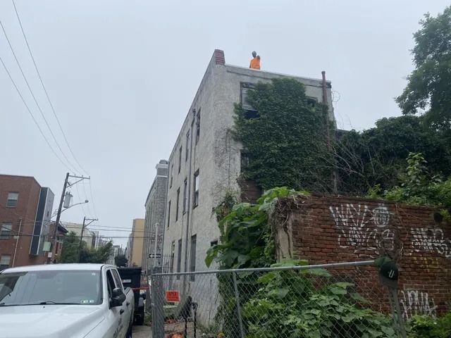 Damaged brick building with ivy, fenced lot, parked car, and worker on the rooftop under a gray sky