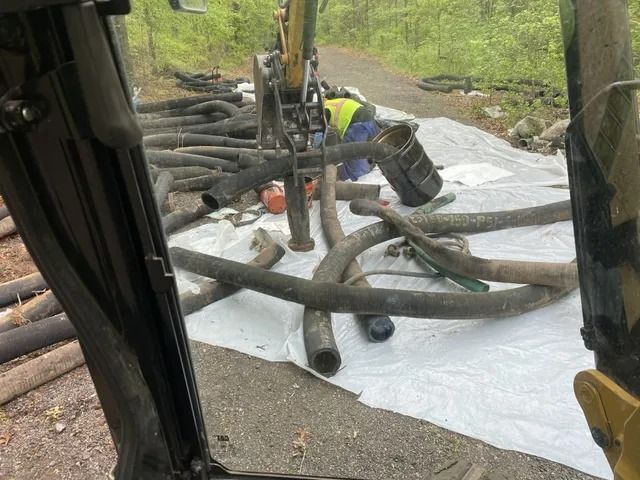 Worker cutting tree branches beside an excavator on a tarp outdoors.