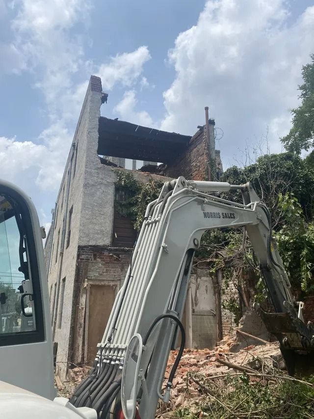 Excavator beside a damaged, partially demolished building with debris and trees around it