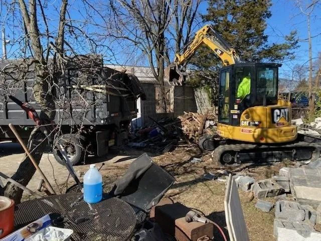 Yellow excavator clearing debris outside a damaged house on a sunny day