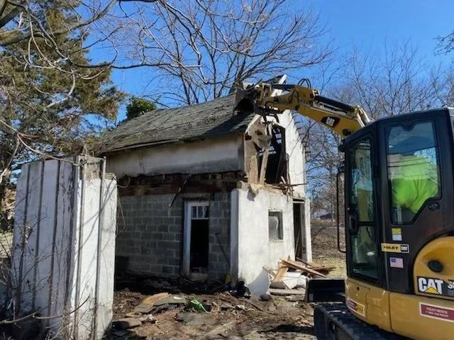 Excavator demolishing a small house with torn roof and white walls under a clear blue sky