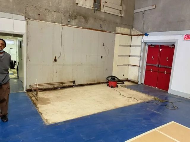 Indoor gym corner with a white wall, red double doors, and a vacuum on the floor.