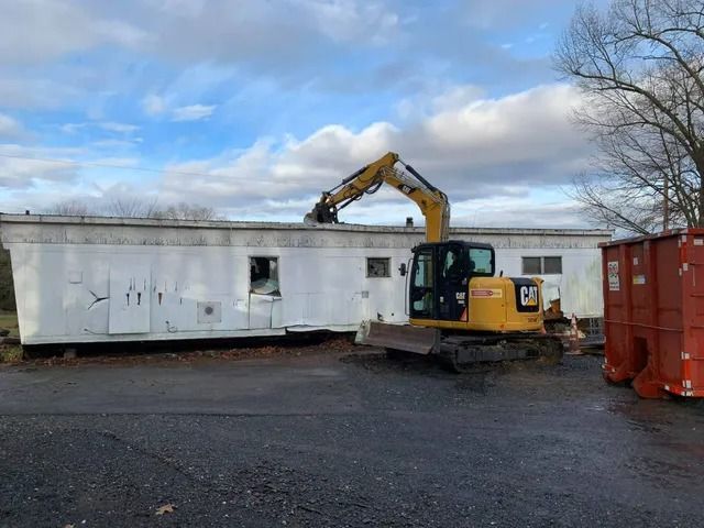 Yellow excavator beside a white portable building and red dumpster in an industrial lot