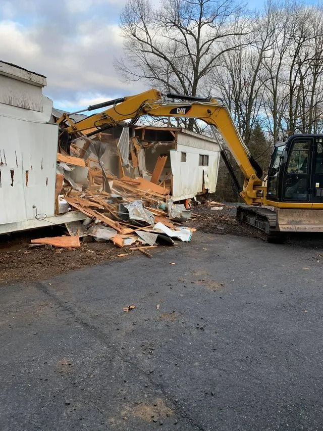 Excavator demolishing a small building, with rubble scattered beside a white structure and a parked machine.