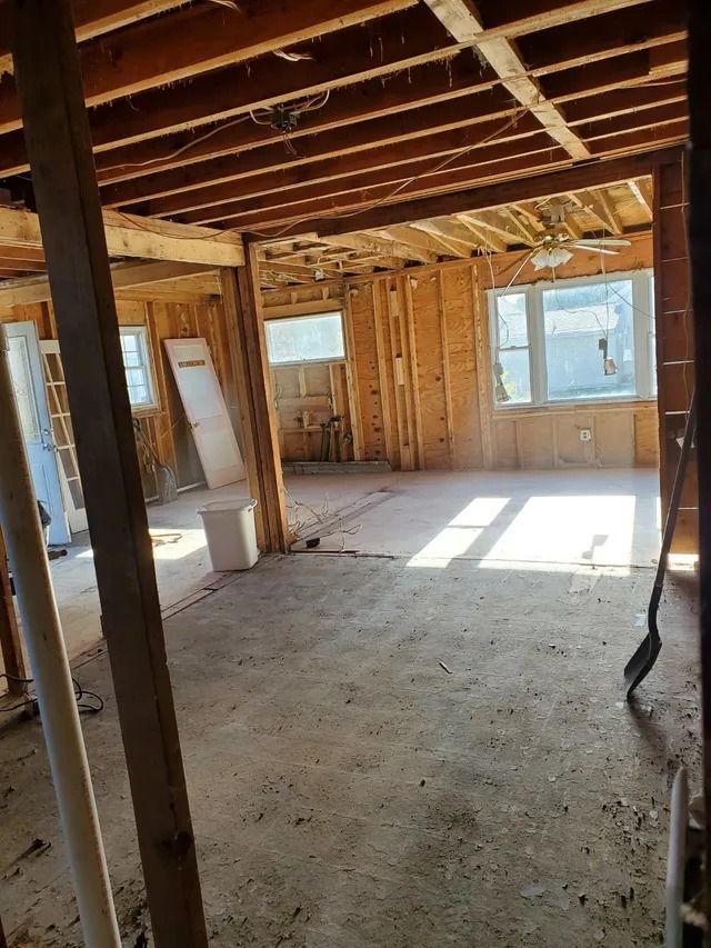 Exposed-framing room under renovation with plywood walls, bare subfloor, and sunlight through windows.