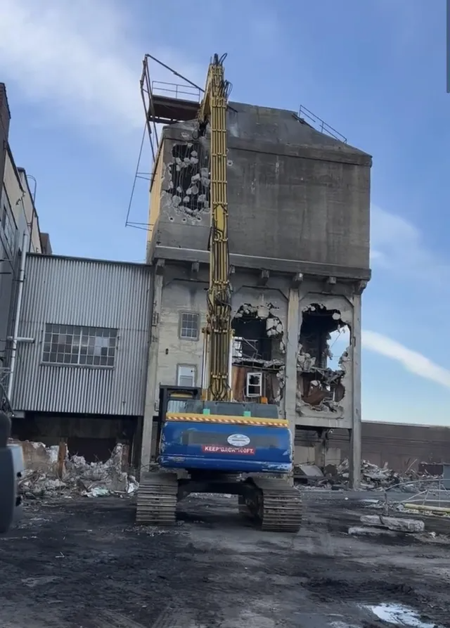 Excavator beside a heavily damaged industrial building, with rubble and debris scattered on the ground.