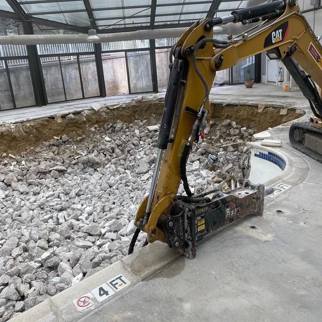 Yellow excavator breaking concrete beside a large indoor excavation pit with rubble and curved edge markings.