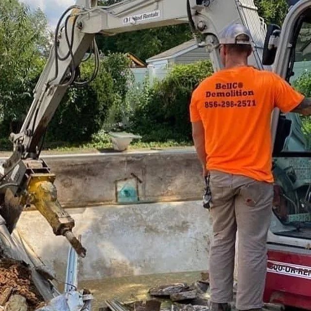 Demolition worker in orange shirt using an excavator to break concrete beside a pool