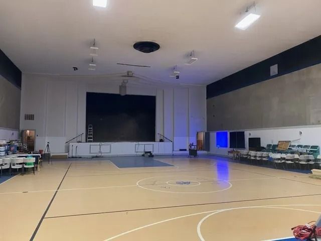 Empty gymnasium with polished floor, stage, wall screens, and rows of chairs along the sides.