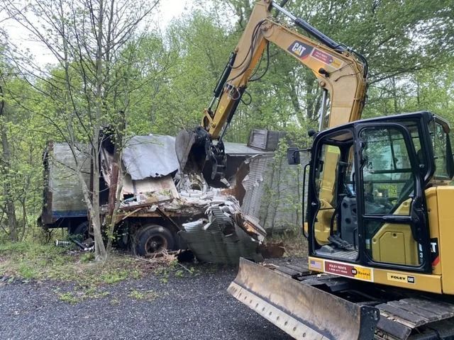 Excavator crushing a burned-out truck in a wooded roadside area