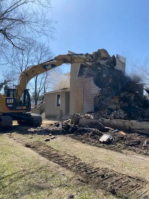 Excavator demolishing a house, with debris piled beside the building on a sunny day.