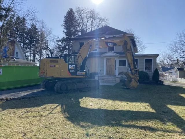 Yellow excavator demolishing a house in a residential yard on a sunny day