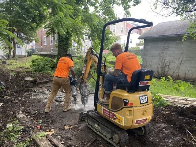 Two workers in orange shirts using construction equipment on a muddy outdoor site beside a house