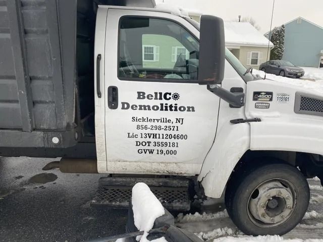 White truck with “Beldi’s Demolition” logo parked in snow, with a dog standing by the door.