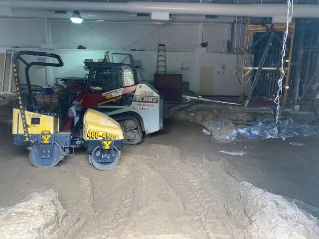 Indoor construction garage with skid steer, road rollers, and equipment on a dirt-covered floor