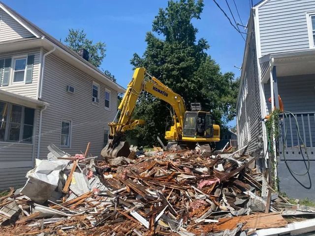 Yellow excavator demolishing a house, with debris piled between neighboring homes.