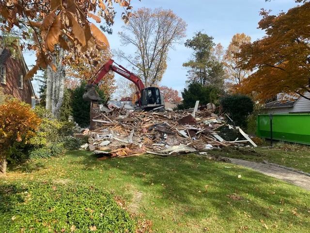 Excavator clearing a large pile of debris in a leafy yard beside a green dumpster