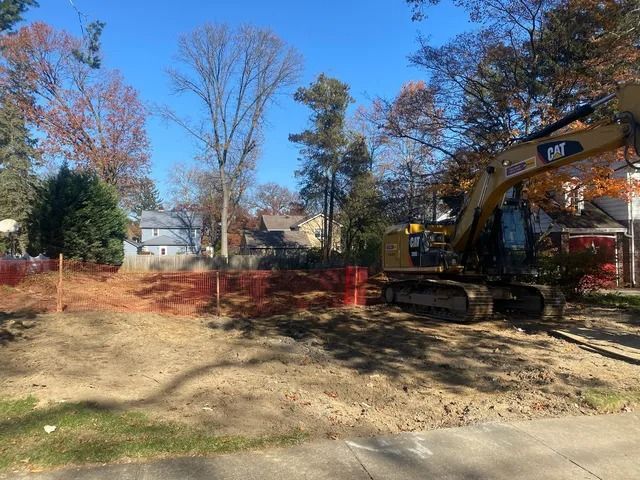 Backhoe excavator clearing dirt in a yard behind orange safety fencing on a sunny day