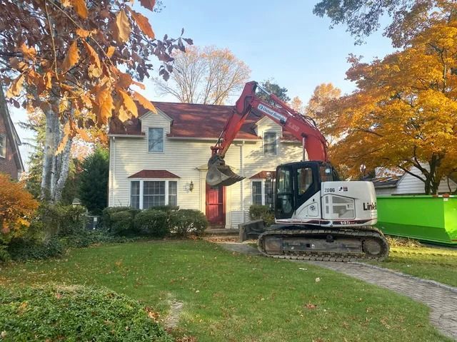 Red excavator beside a two-story house, clearing a front lawn in autumn.