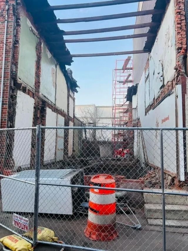 Construction site with damaged walls, exposed roof beams, and a chain-link fence in the foreground.