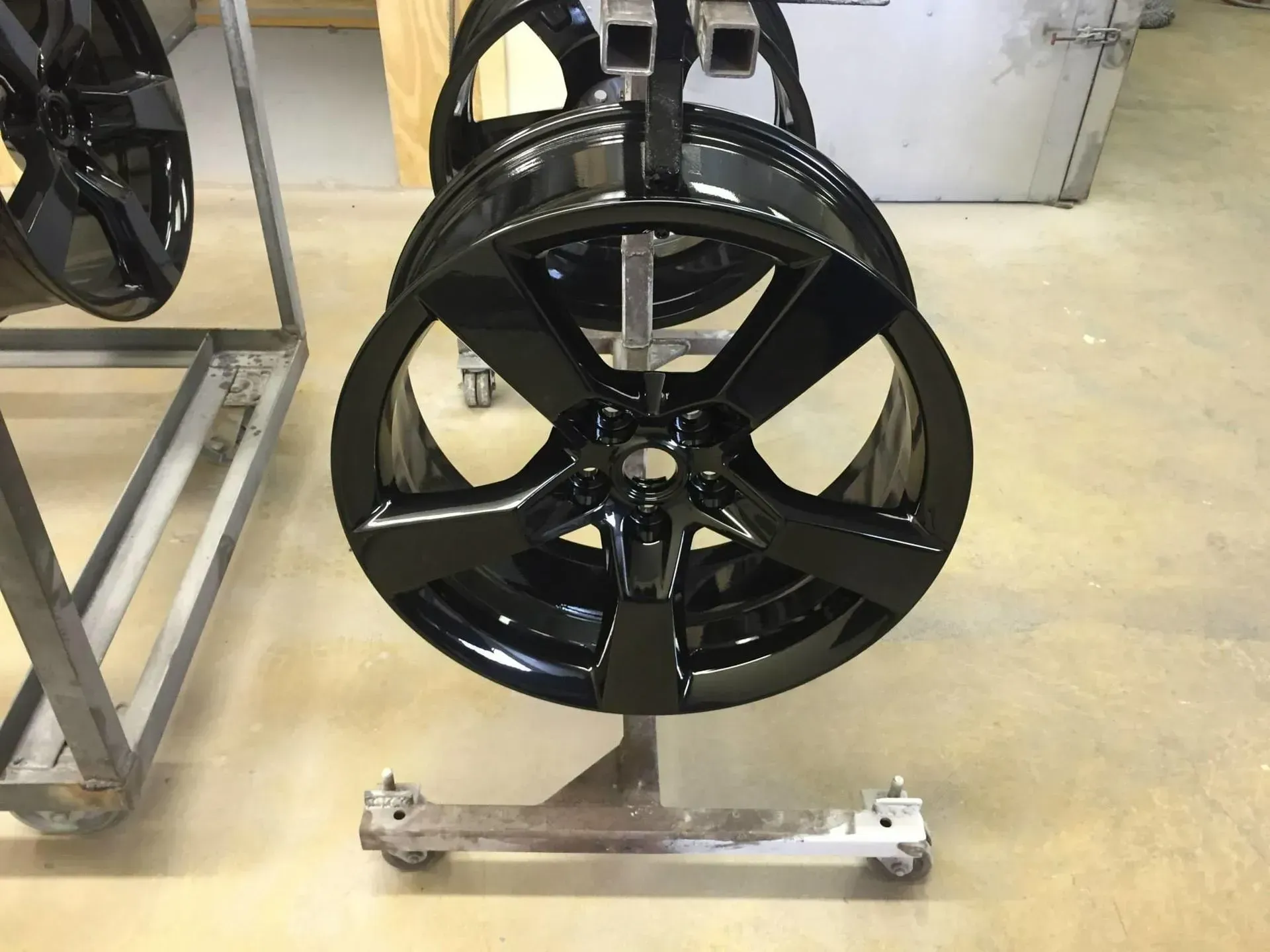 Glossy black five-spoke automotive wheels mounted on metal racks in a workshop.