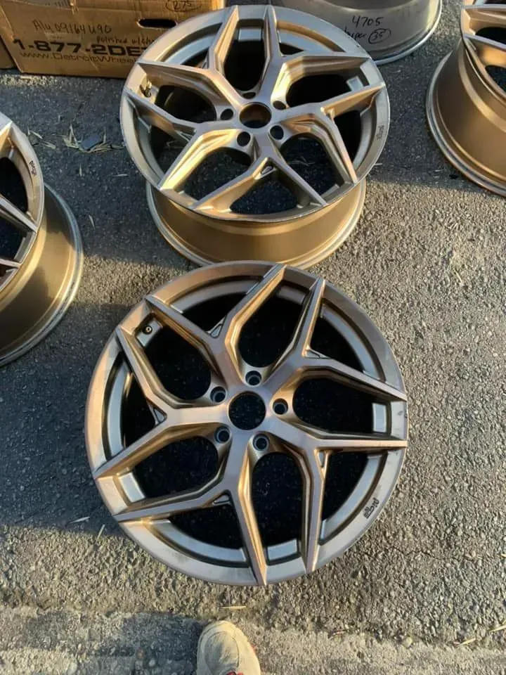 Two bronze-colored, five-spoke automotive alloy wheels resting on pavement near a cardboard box.