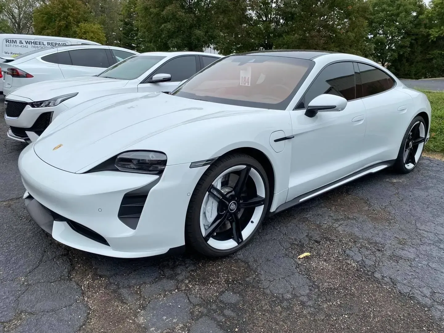 A white Porsche Taycan parked on an asphalt lot, with a white sedan visible in the background.