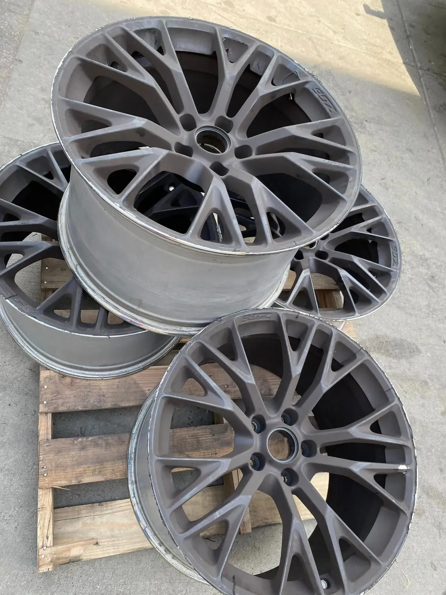 Four dark gray, multi-spoke alloy car wheels arranged on a wooden pallet outdoors on concrete.