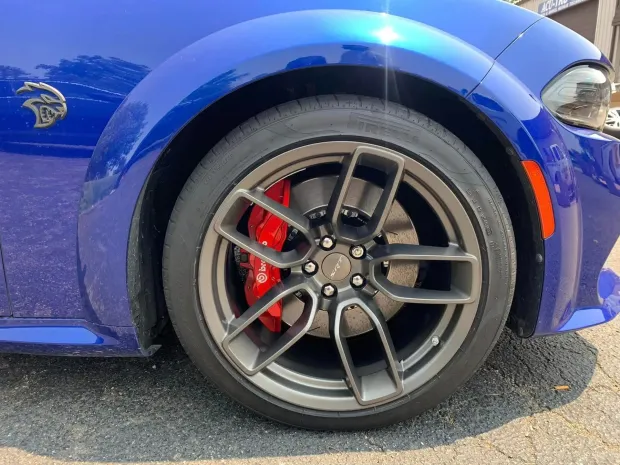 A close-up of a blue Dodge Challenger's front wheel, featuring a grey alloy rim, red Brembo brake caliper, and Hellcat badge.