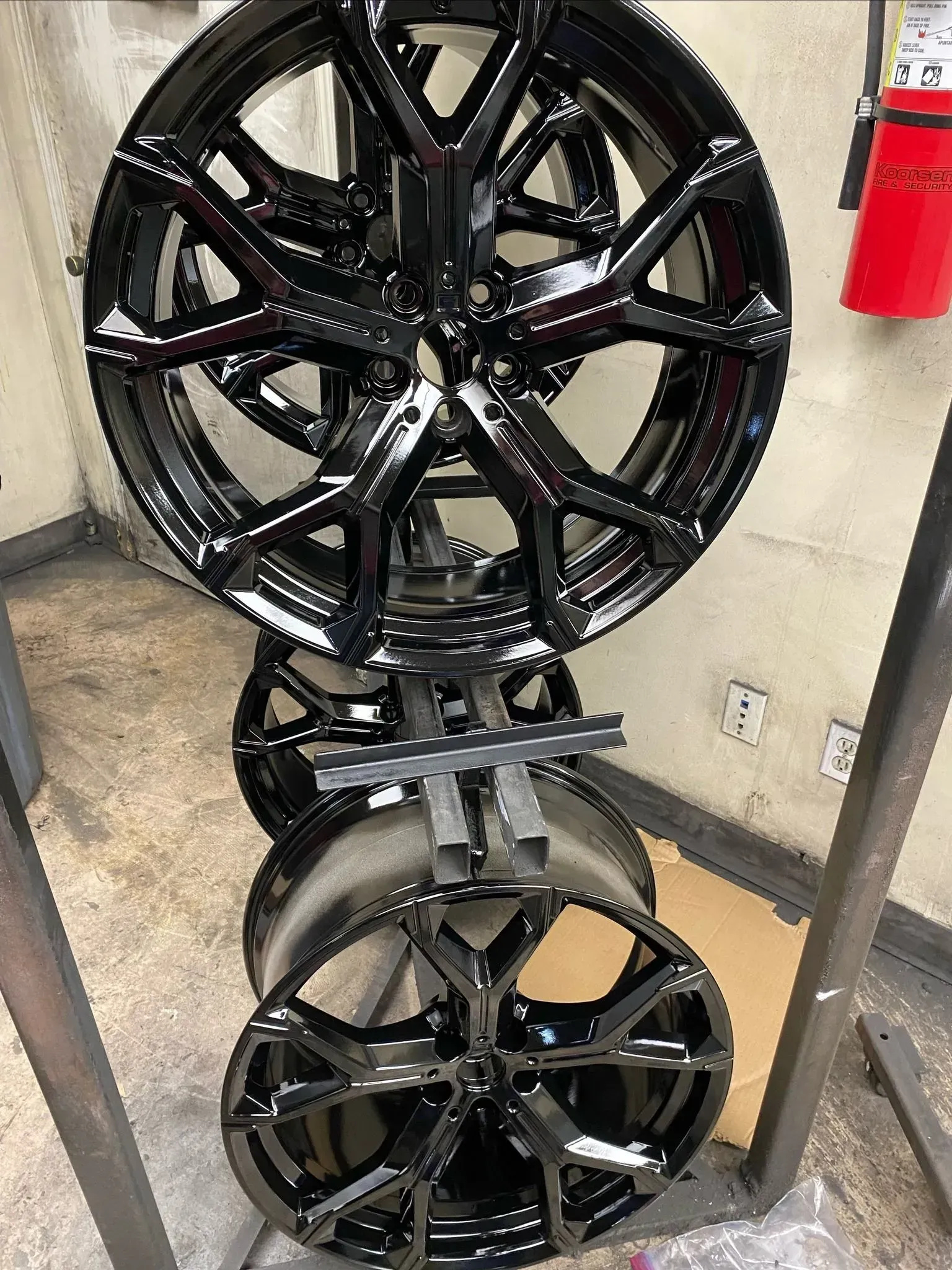 A vertical metal rack holds two glossy black, multi-spoke automotive wheels in a workshop setting.