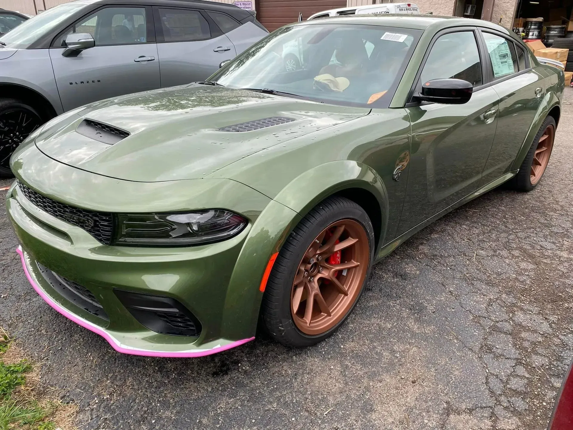 Olive green Dodge Charger widebody sedan with bronze wheels and a pink front splitter edge, parked on an asphalt lot.
