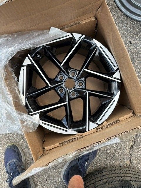 Black-and-silver alloy wheel rim in a cardboard box, viewed from above on pavement.