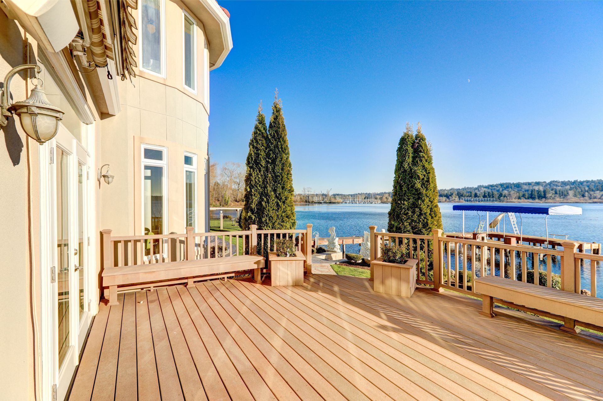 Sunlit seaside deck beside a cream building, with wooden railing, potted cypress trees, and ocean view