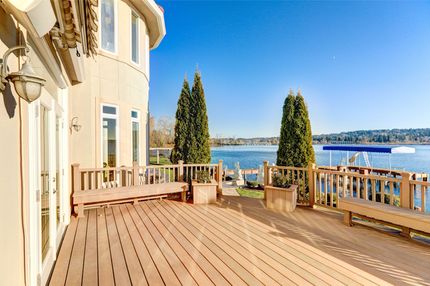 Sunlit seaside deck beside a cream building, with wooden railing, potted cypress trees, and ocean view