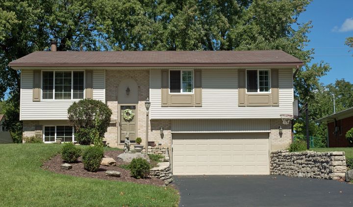 Two-story beige house with attached garage, stone retaining wall, and front lawn under trees