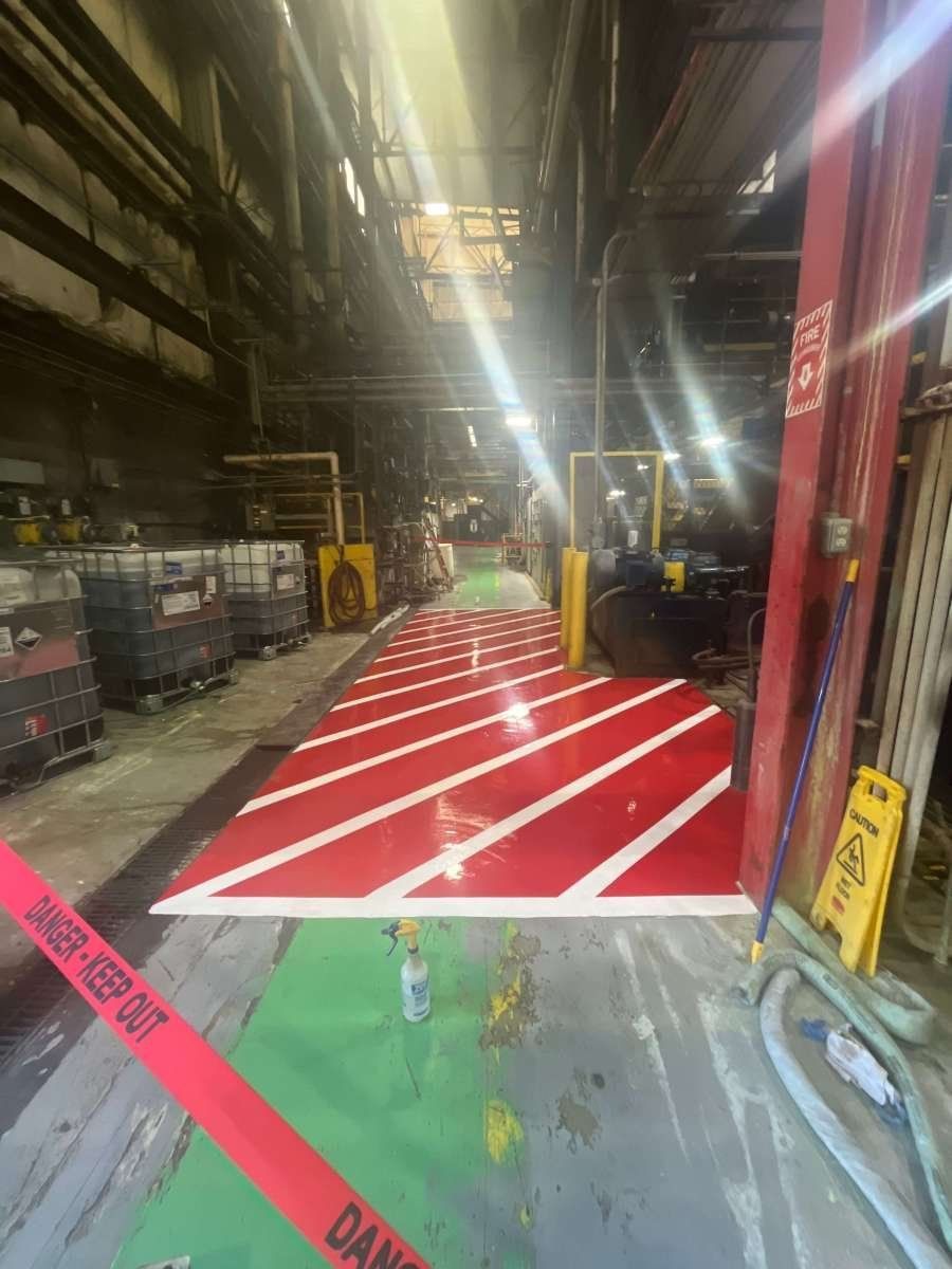 A freshly painted red and white striped safety floor marking in an industrial warehouse, cordoned off with caution tape.