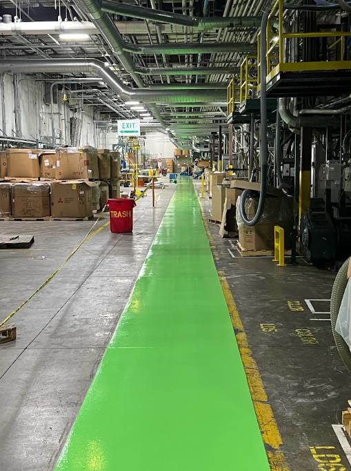 Industrial warehouse aisle with bright green floor path, stacked boxes, and machinery on both sides.