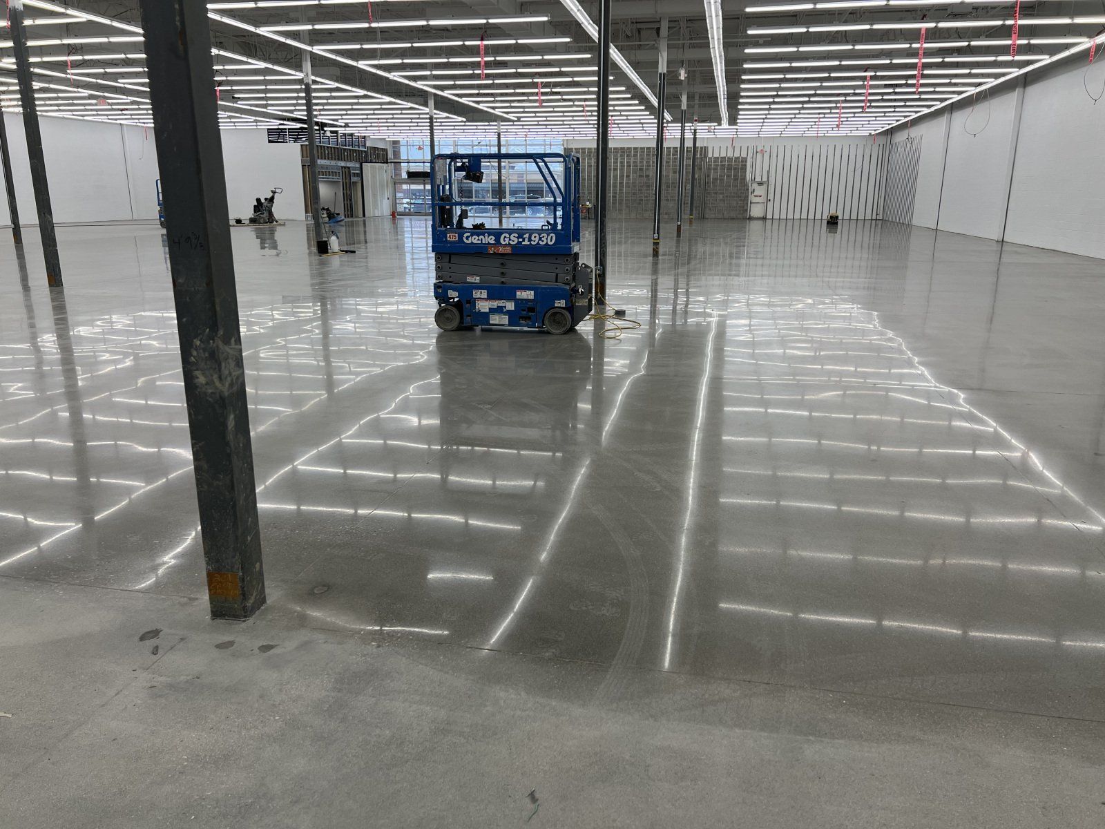 A blue scissor lift sits in the center of a large, empty commercial space with a polished, reflective concrete floor.
